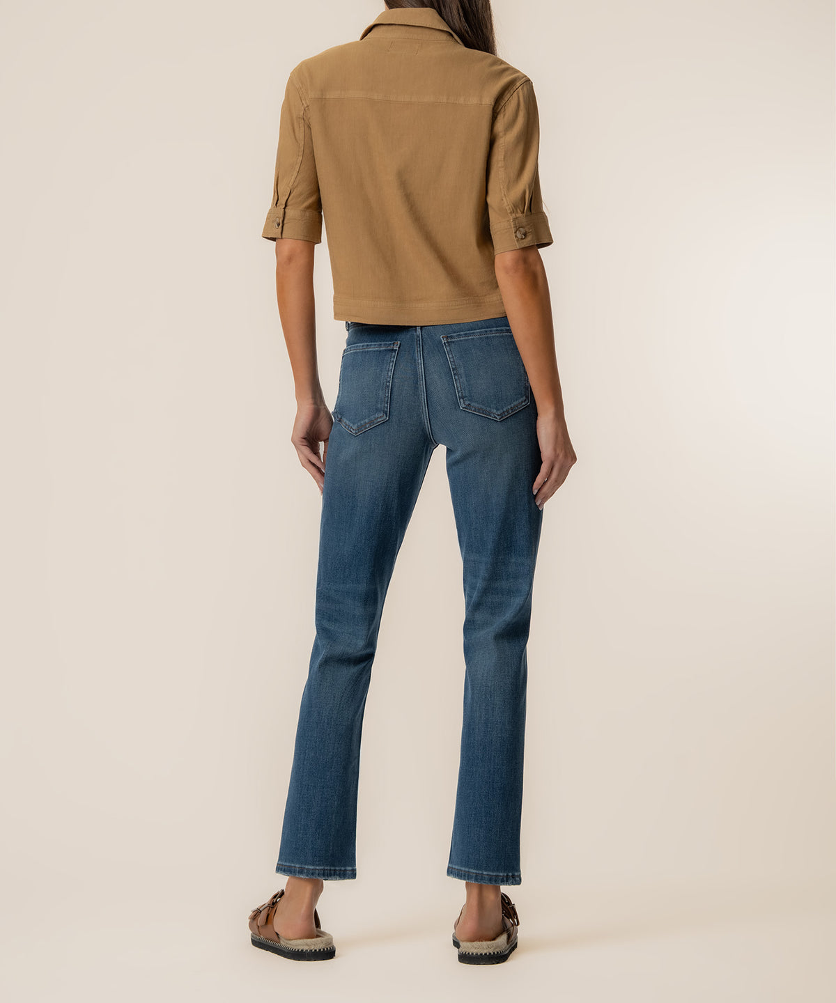 Woman wearing a tan linen jacket with half sleeves and blue denim jeans, viewed from behind.