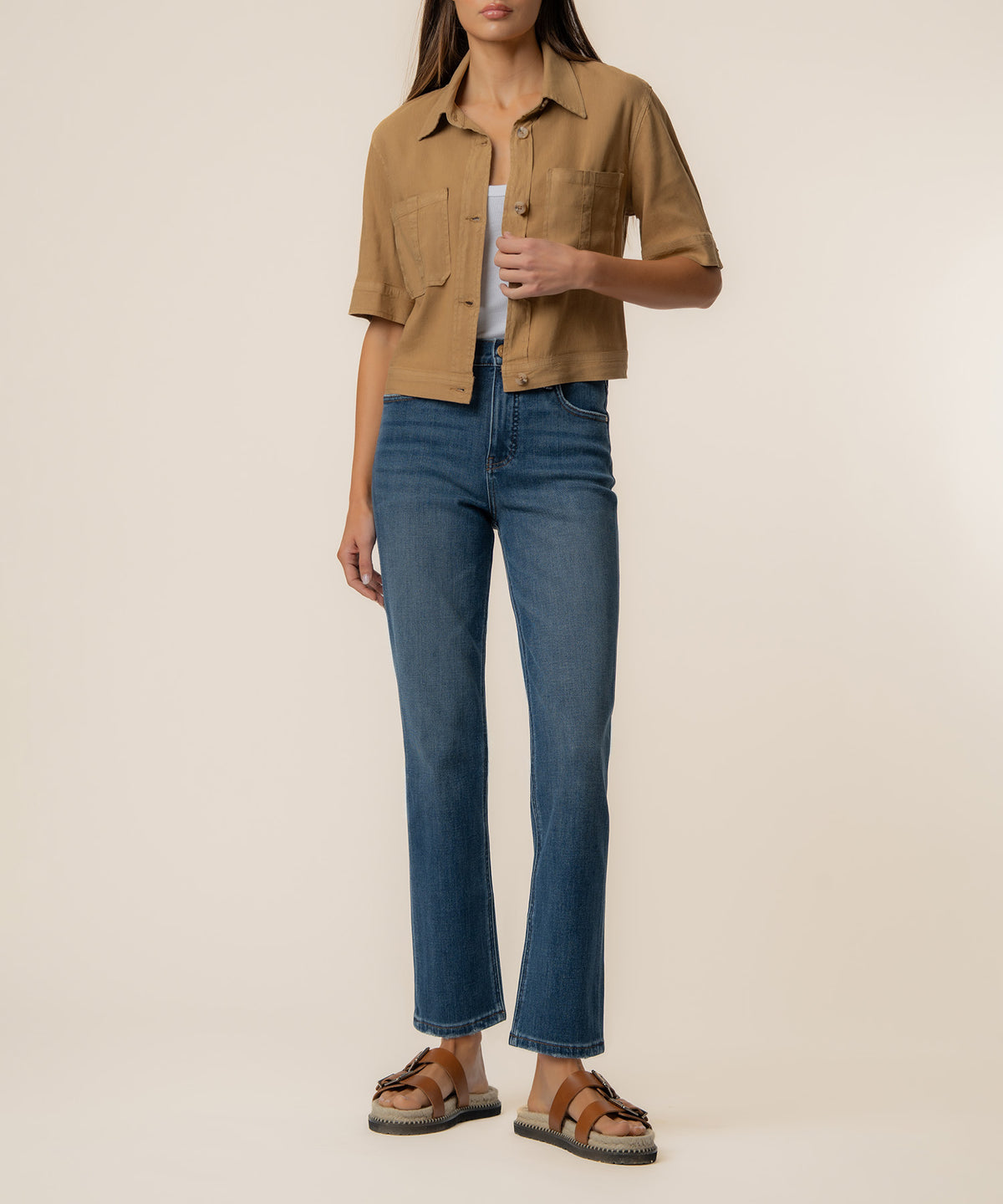 Woman wearing a tan linen jacket with chest pockets and short sleeves, styled with blue jeans and brown sandals.