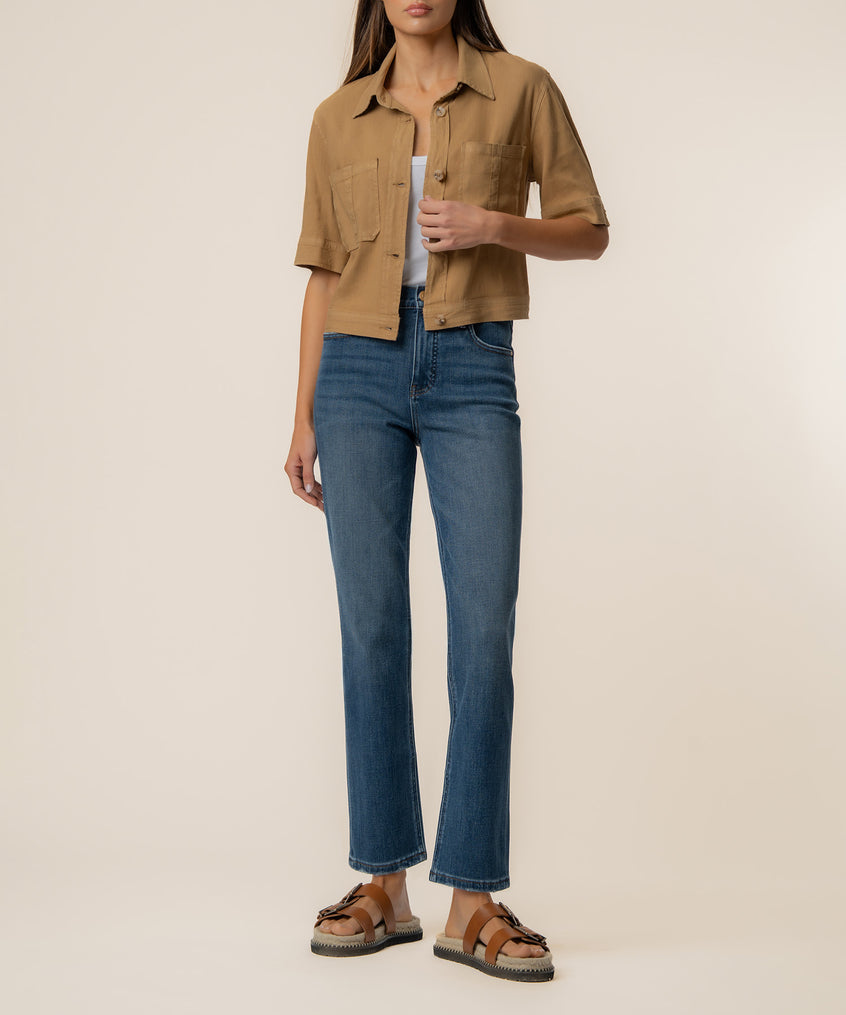 Woman wearing a tan linen jacket with chest pockets and short sleeves, styled with blue jeans and brown sandals.
