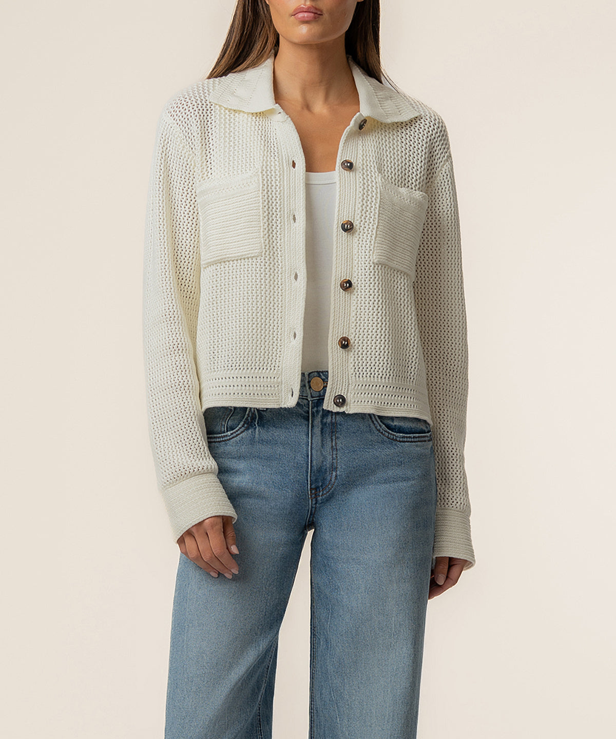 Cream knit sweater jacket with textured panels, collar, and brown button front closure worn over white top and blue jeans.