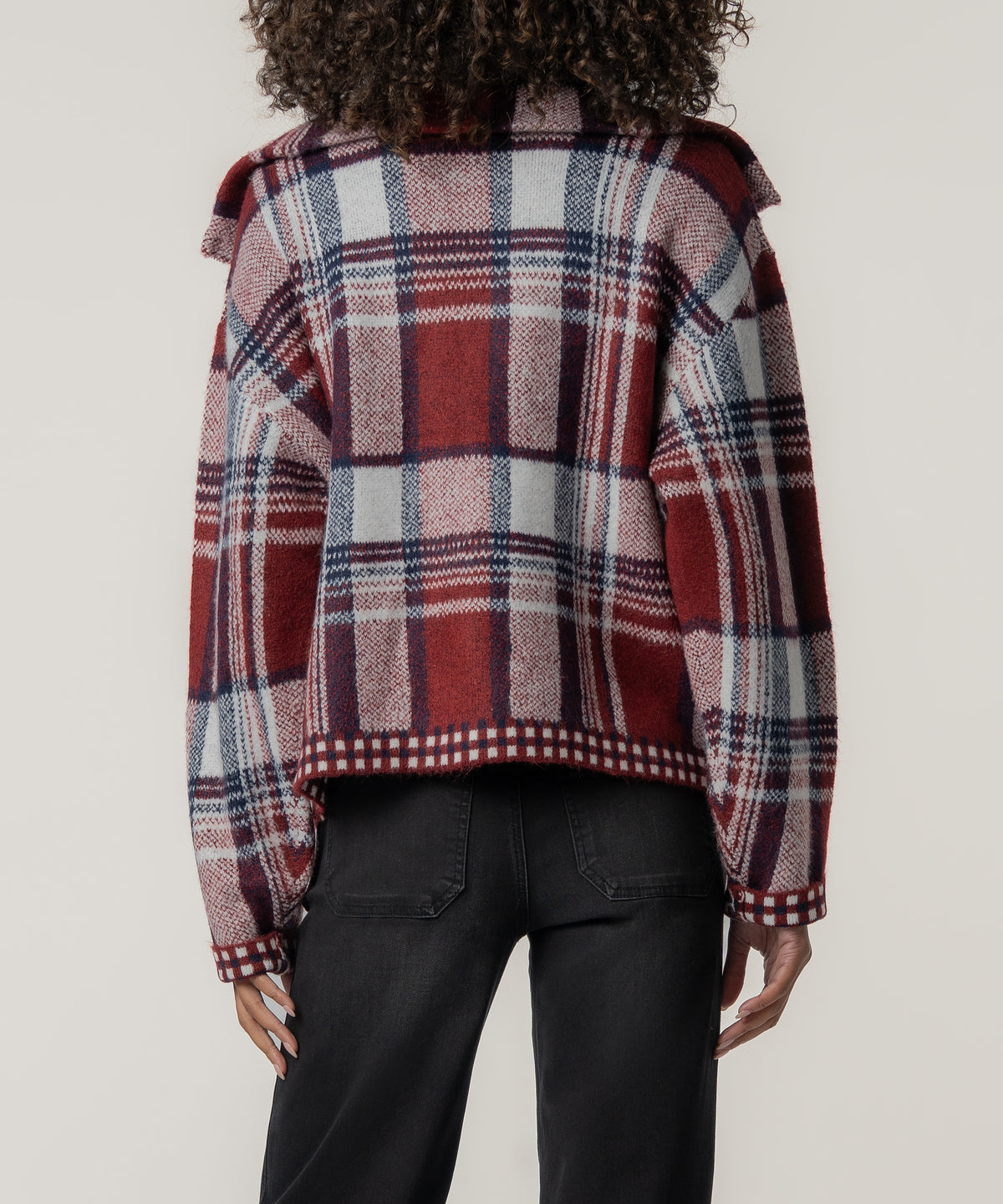 Back view of a person wearing a cropped red, navy, and white plaid knit jacket with checkered trim and black jeans.