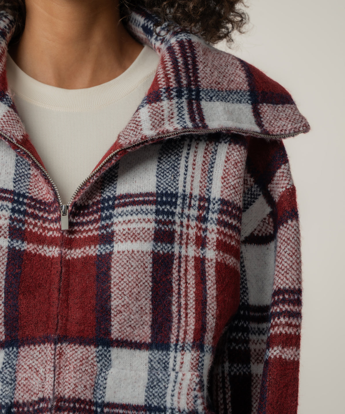 Close-up of a person wearing a red, navy and white plaid knit jacket with a wide collar, silver zipper and cream top.