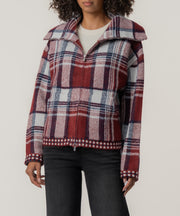 A person wears a zip-front plaid knit jacket in brick red, navy, and white with a wide collar, over black jeans.