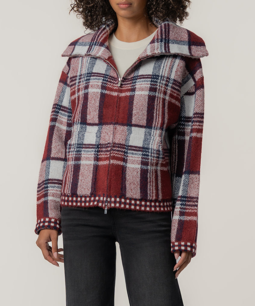 A person wears a zip-front plaid knit jacket in brick red, navy, and white with a wide collar, over black jeans.