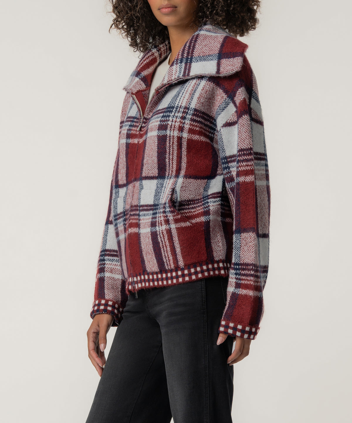 A woman wears a zip-front plaid knit jacket in brick red, navy, and light blue with checkered trim, styled with black jeans.