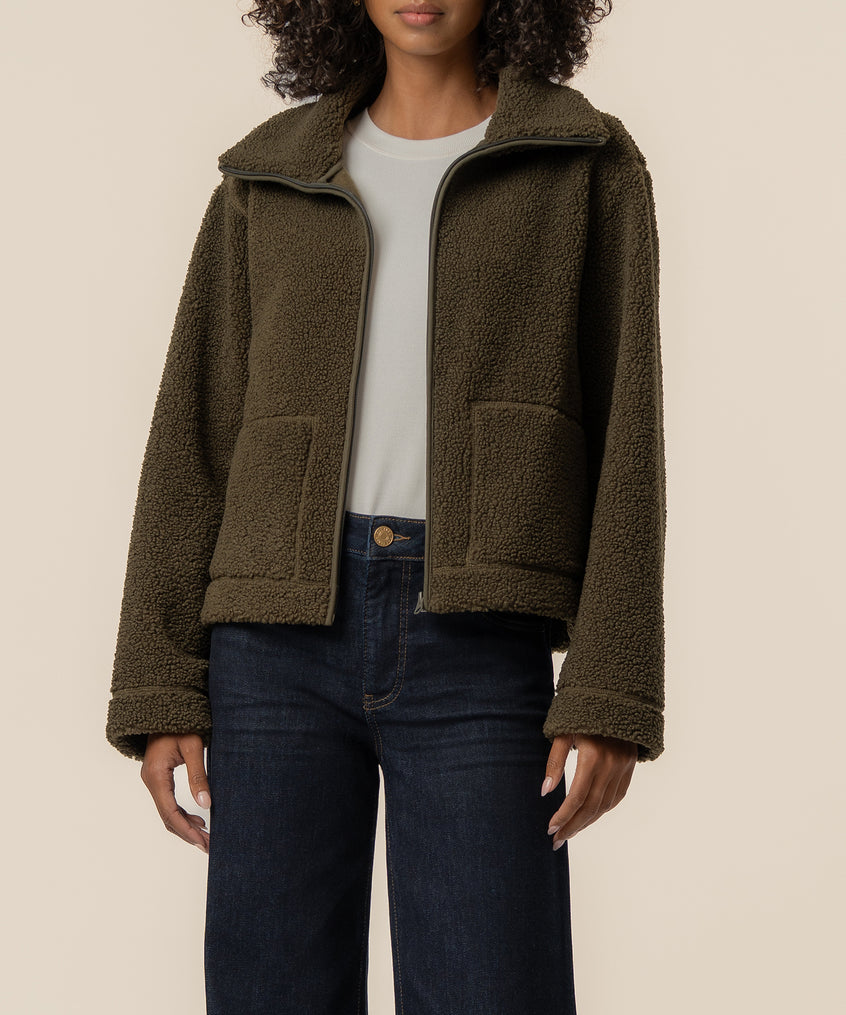 Olive faux shearling jacket with wide collar, full-zip front, and patch pockets worn over white tee and dark jeans.