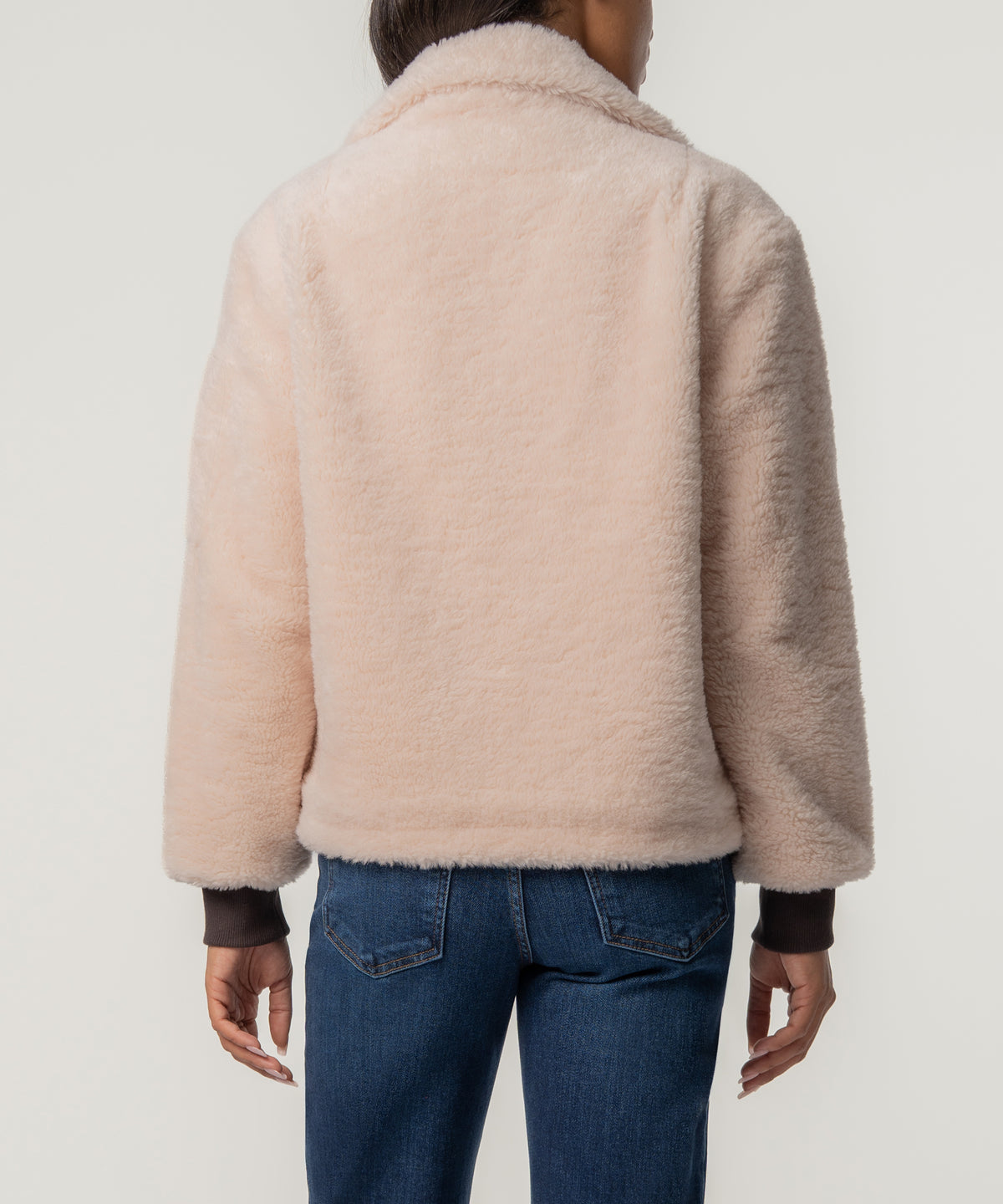 Back view of a person wearing a cropped cream faux sherpa jacket with dark ribbed cuffs and blue jeans.