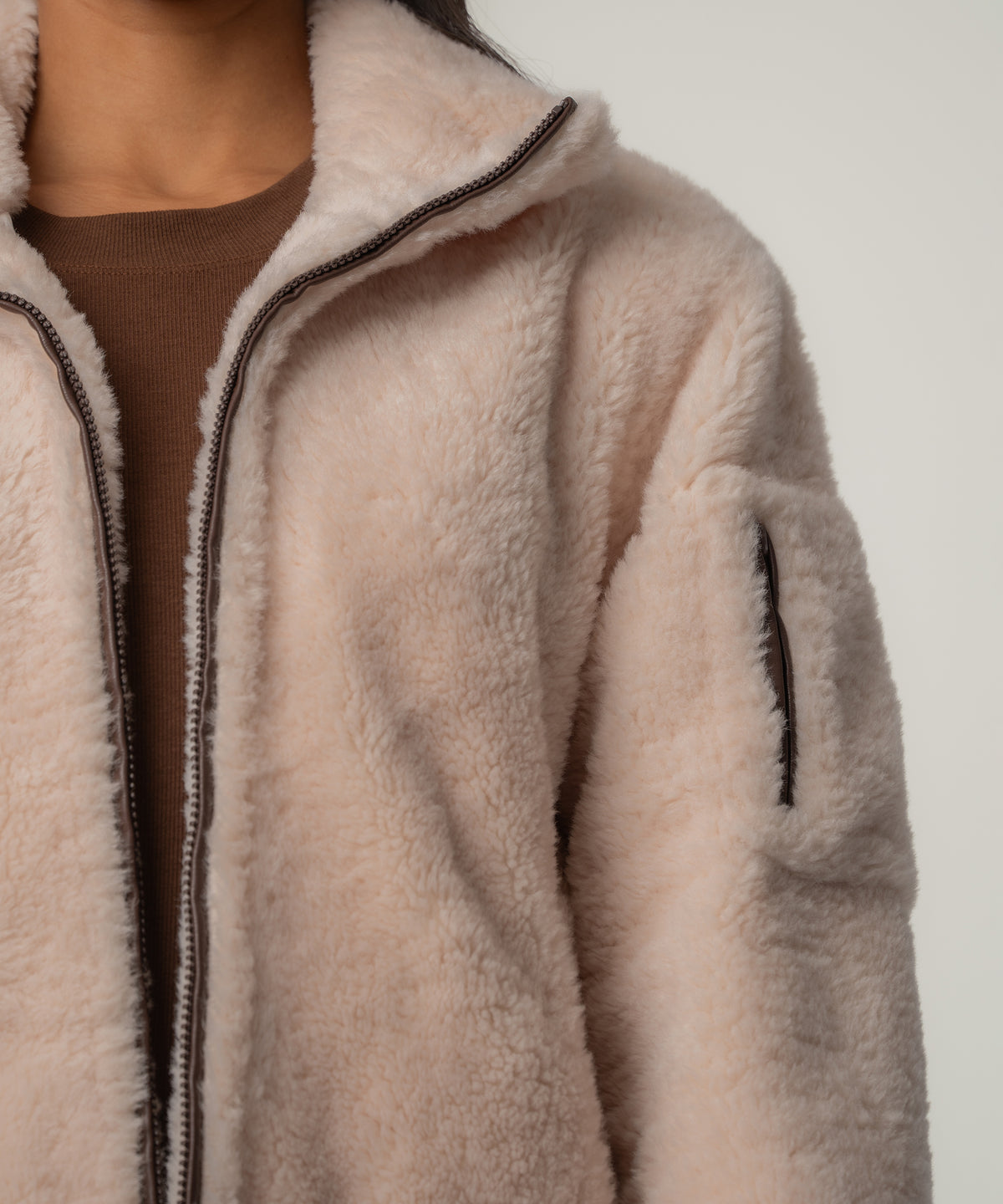 A cream faux sherpa jacket with a brown zipper and sleeve pocket is worn open over a brown top.