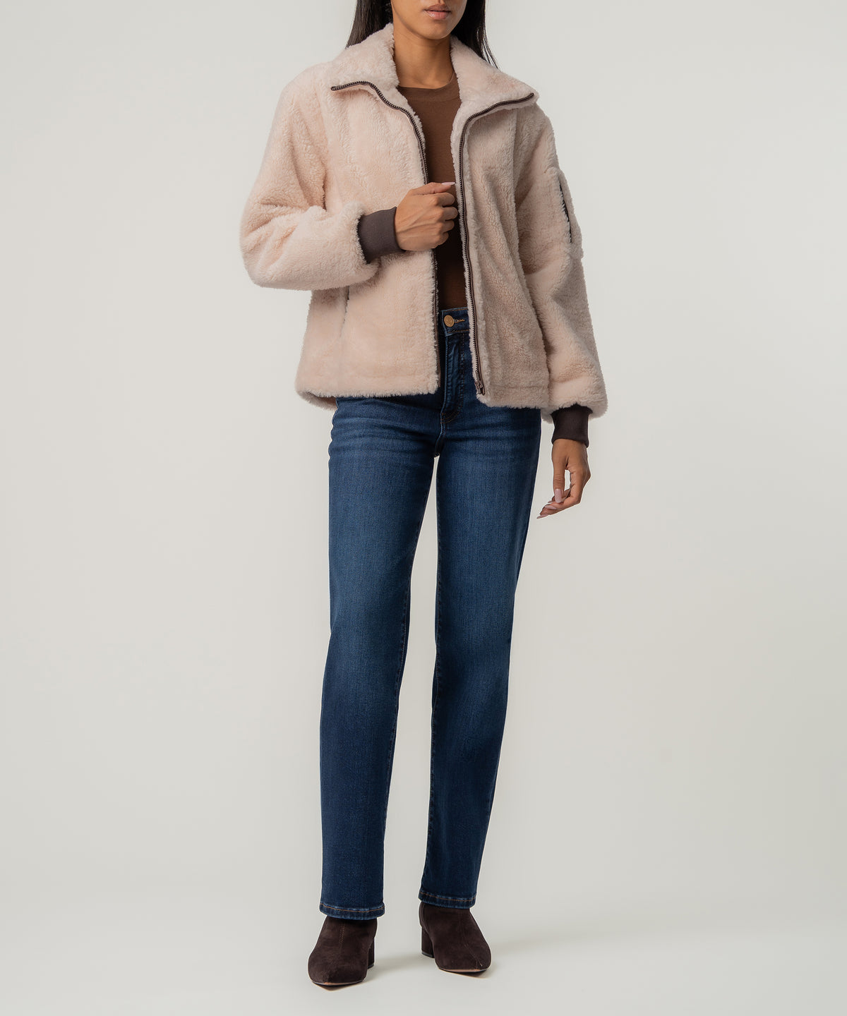 A woman wears a cream faux sherpa zip jacket with brown ribbed cuffs, a brown top, dark blue jeans, and brown ankle boots.