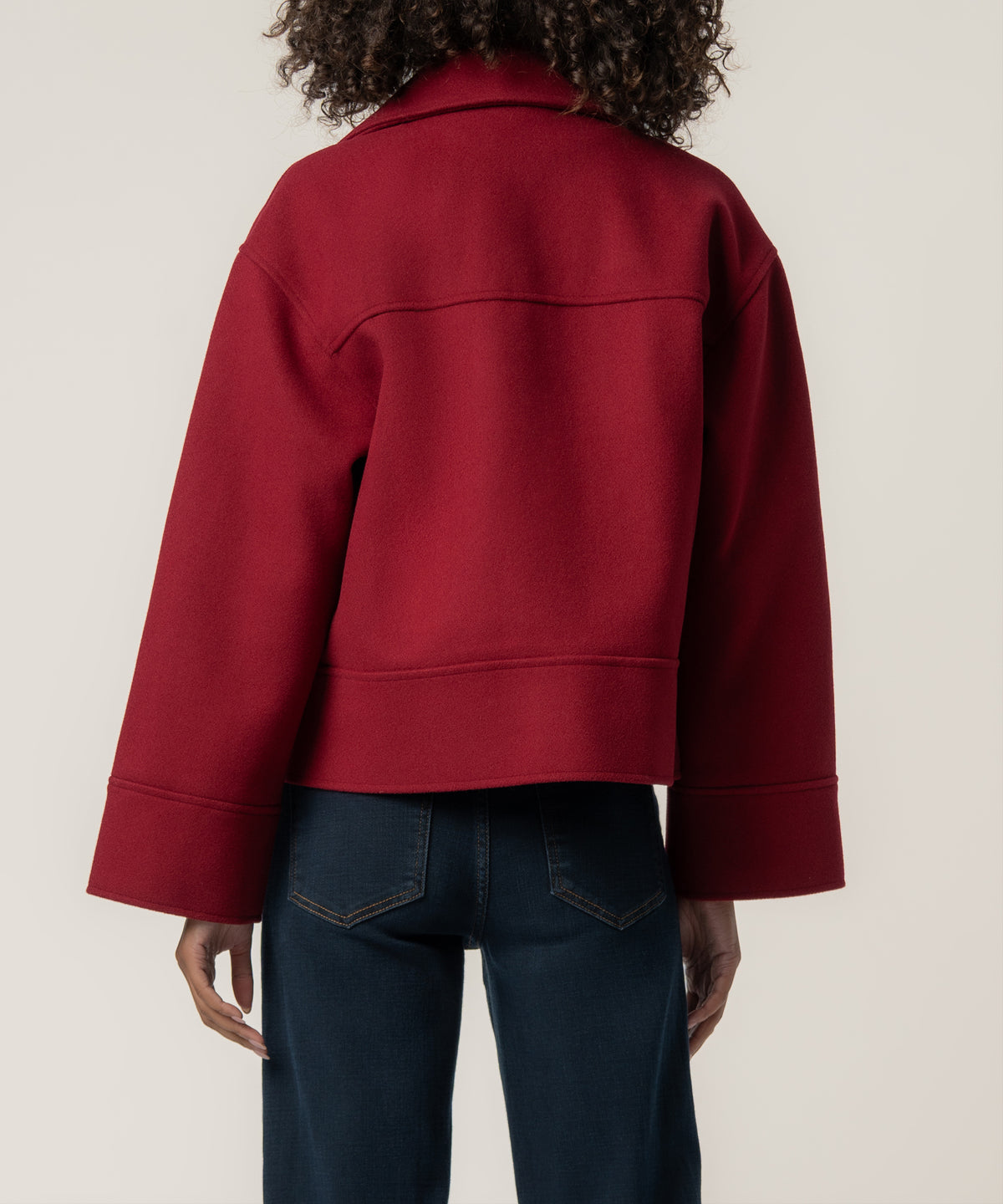 Back view of a person wearing a cropped crimson wool jacket with wide sleeves and dark blue jeans.