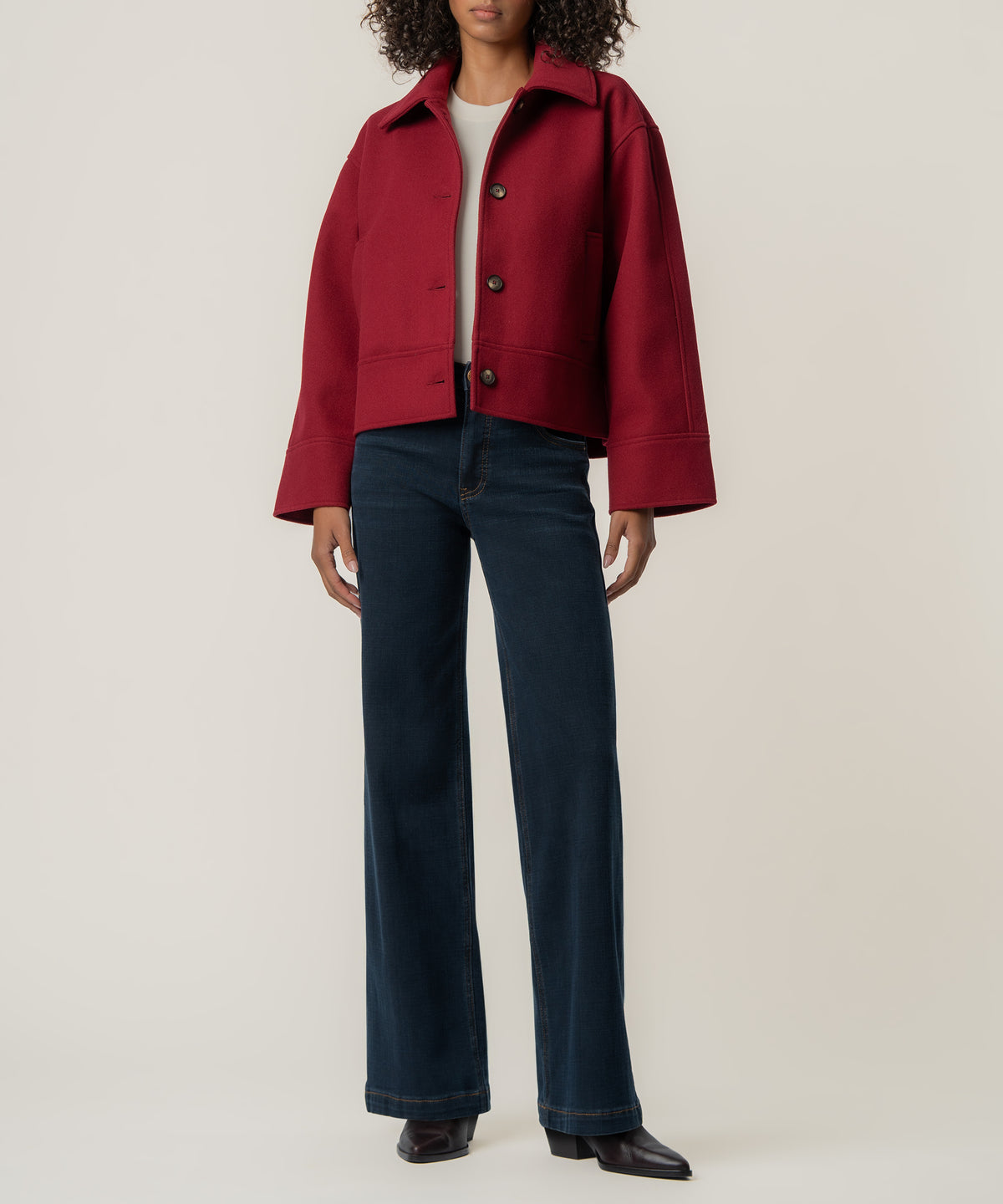 A person wears a cropped crimson jacket with buttons and wide sleeves over a cream top, dark flared jeans, and black boots.