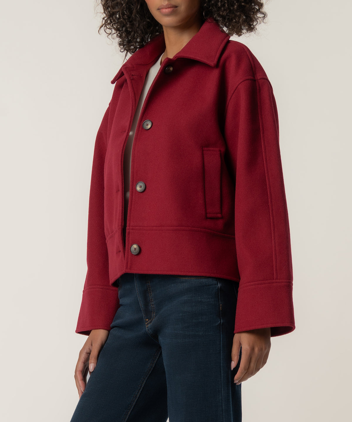 A woman wears a cropped crimson wool jacket with a pointed collar, brown buttons, and wide cuffs over dark blue jeans.