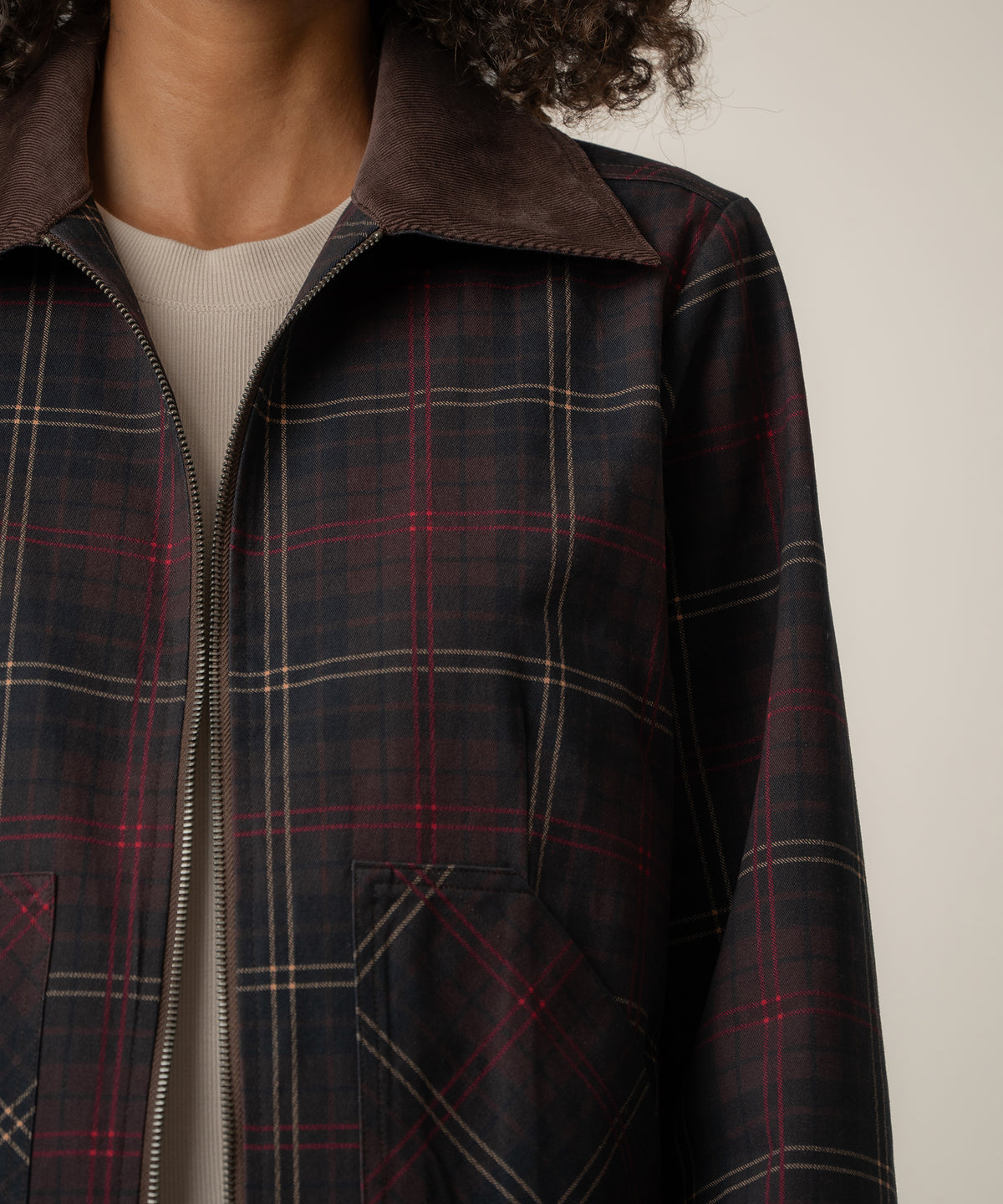 A person wears an unzipped dark plaid jacket with red and tan lines, a brown corduroy collar, and a cream top.