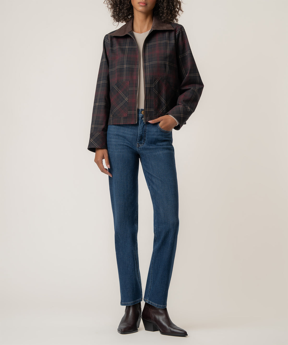 A woman wears a cropped dark plaid jacket with patch pockets, a beige top, blue straight-leg jeans, and dark brown ankle boots.