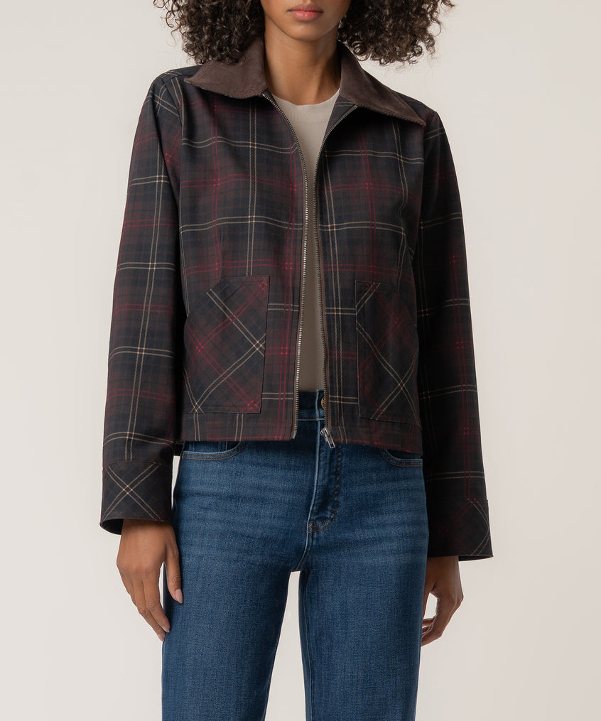 A woman wears a dark plaid zip-front jacket with a brown corduroy collar and patch pockets over blue jeans.