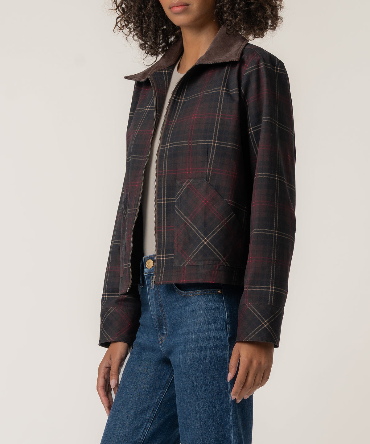 A woman wears a cropped zip-up plaid jacket with a brown corduroy collar over a light top and blue jeans.