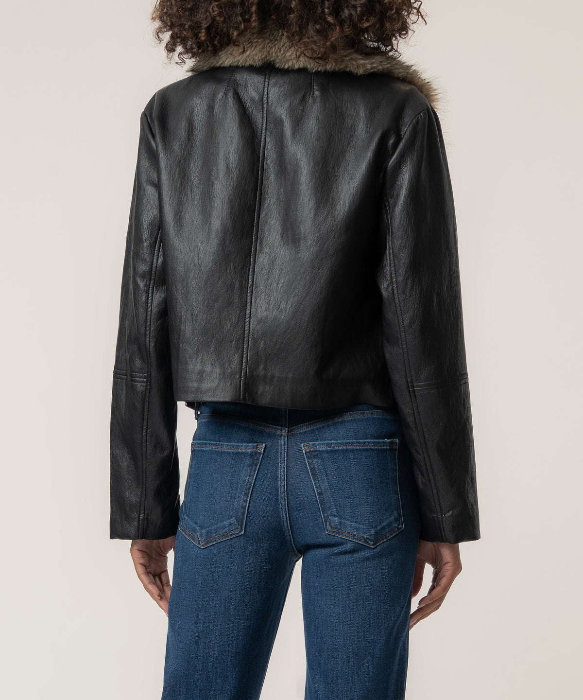 Back view of a person wearing a cropped black faux leather jacket with a fur collar and blue high-waisted jeans.