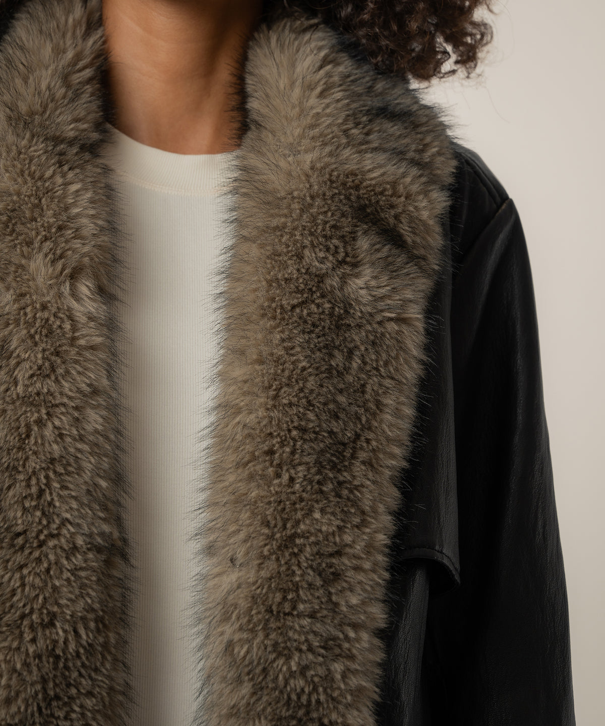 Close-up of a black faux leather jacket with a thick taupe faux fur collar, worn over a cream ribbed top.