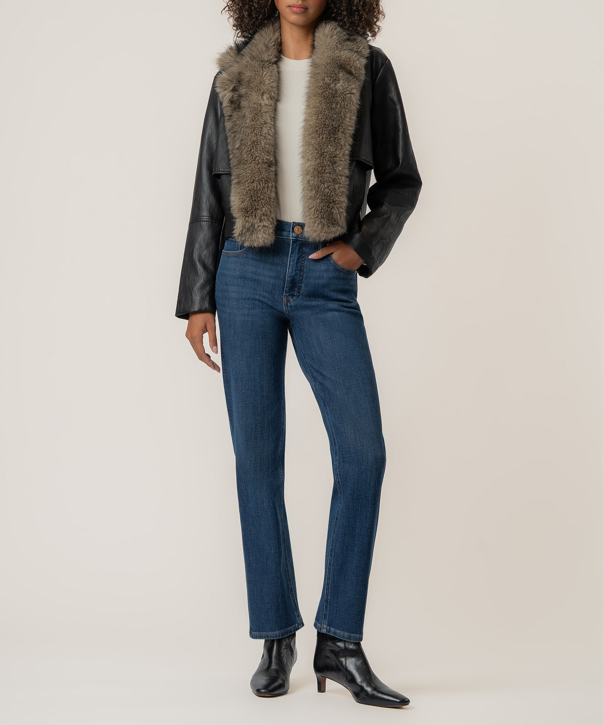A woman wears a cropped black faux leather jacket with a taupe faux fur collar, blue jeans, and black ankle boots.