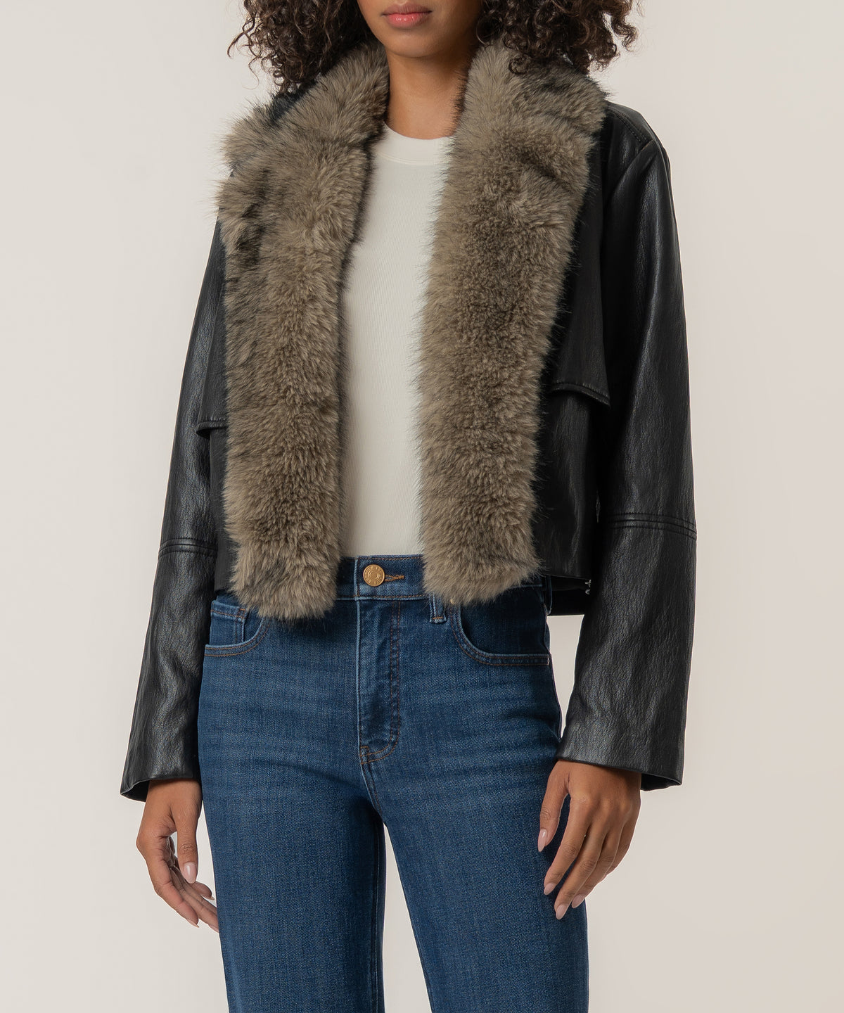A woman wears a cropped black faux leather jacket with a thick taupe fur collar over a white top and blue jeans.