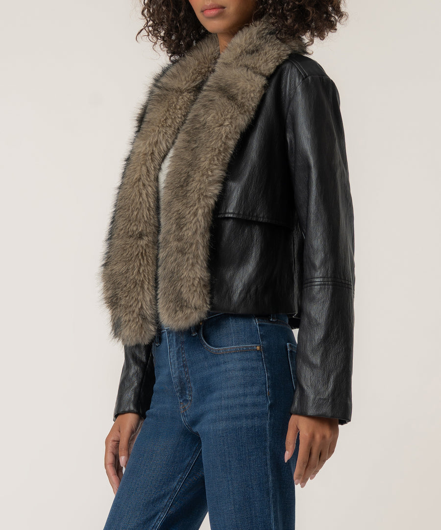 A woman wears a cropped black faux leather jacket with a thick taupe fur collar, paired with blue jeans.