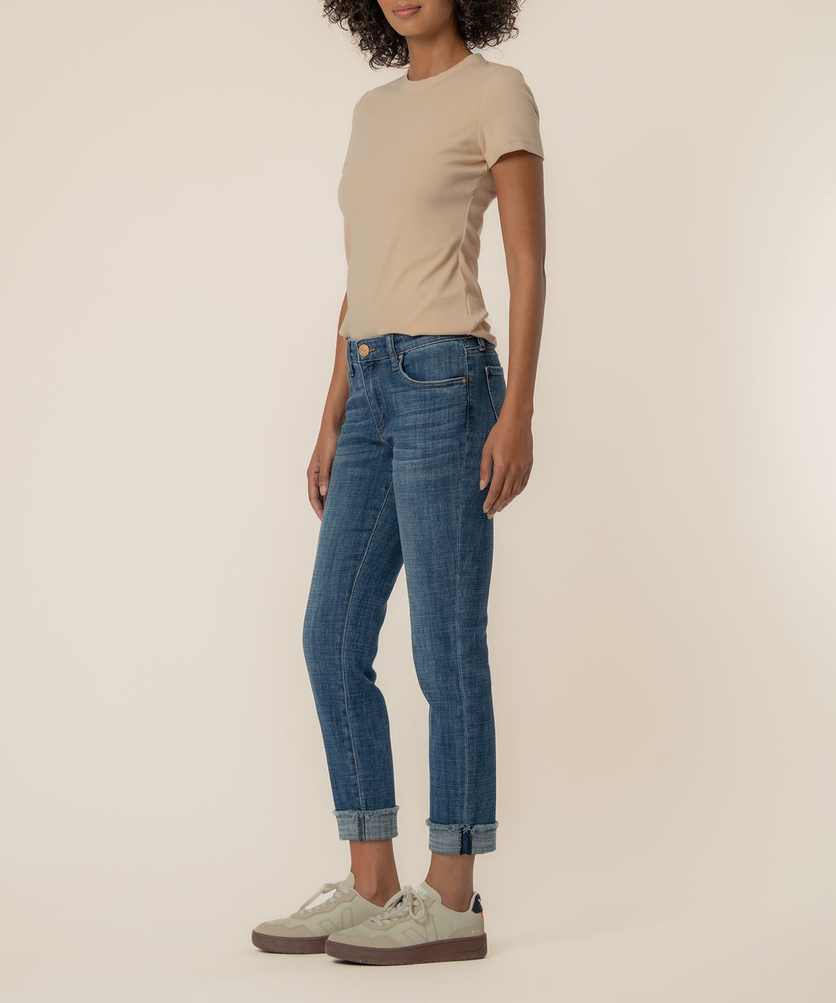 Woman wearing beige t-shirt and mid-rise blue denim crop jeans with rolled cuffs and cream sneakers.