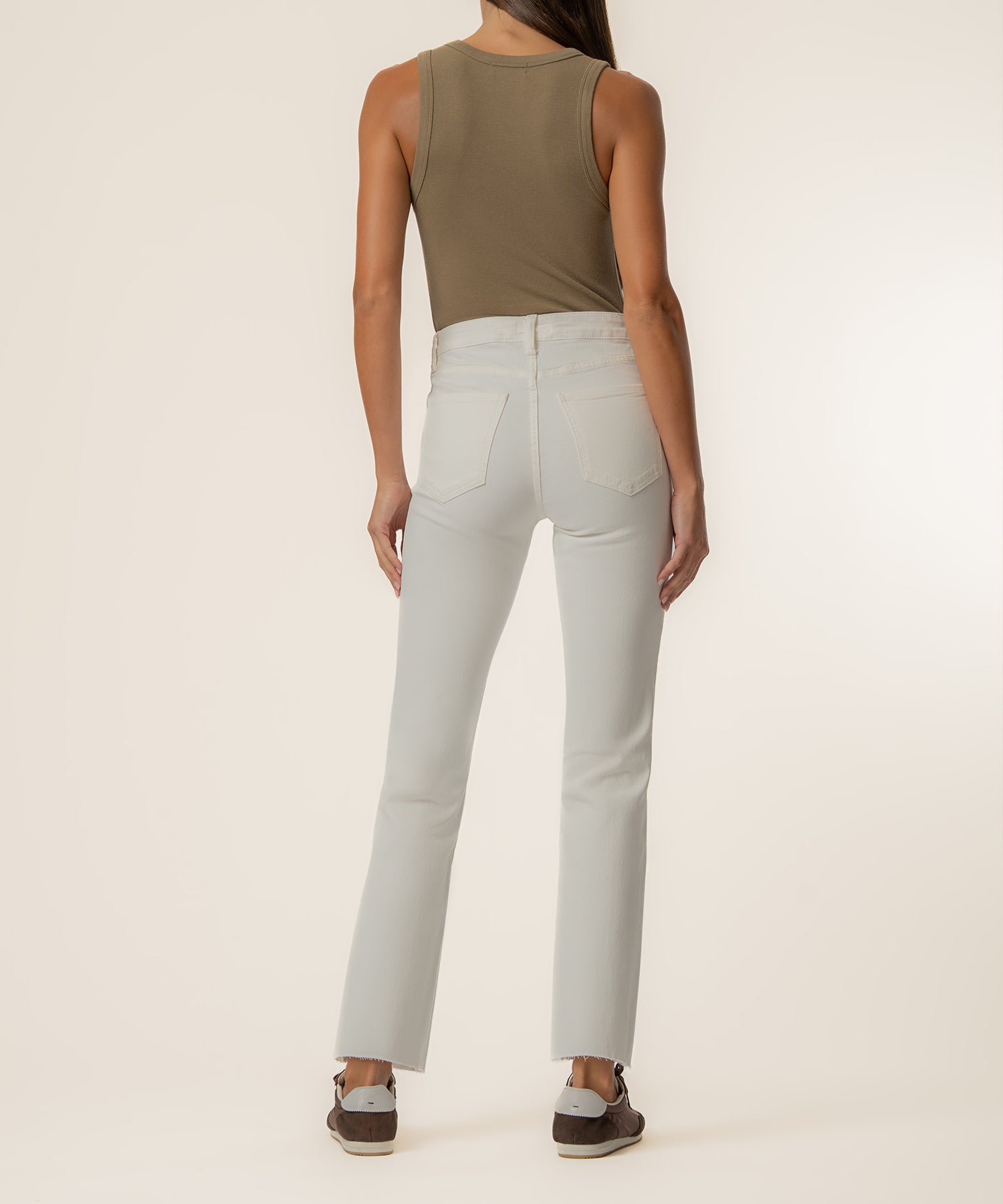 Woman wearing ecru high-rise ankle flare jeans with distressed hem and brown sleeveless top, shown from back.