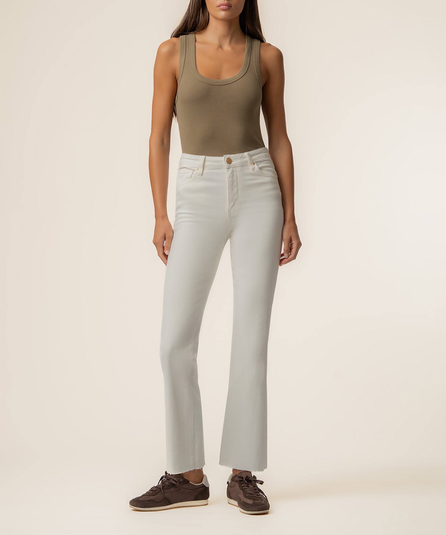 Woman wearing high rise ecru ankle flare jeans with a tan sleeveless top and brown shoes.