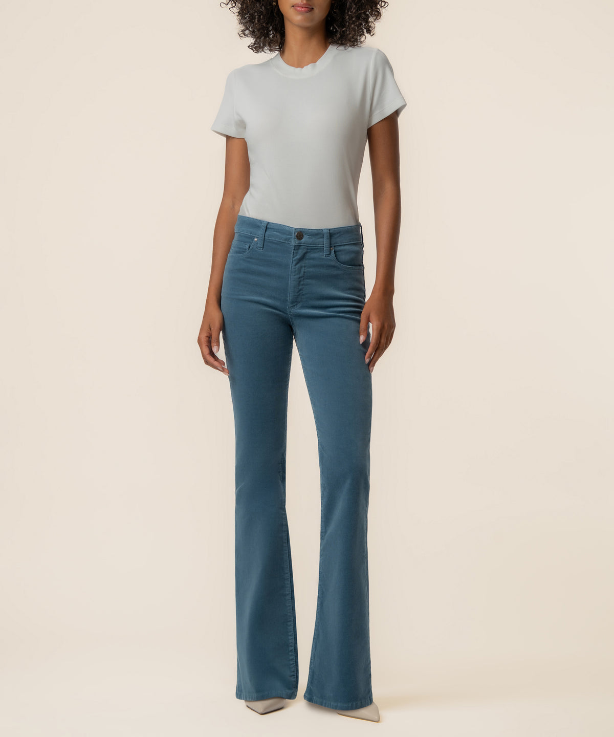 A woman stands against a cream background wearing blue high-rise flare corduroy pants, a pale short-sleeve top, and light pointed shoes.