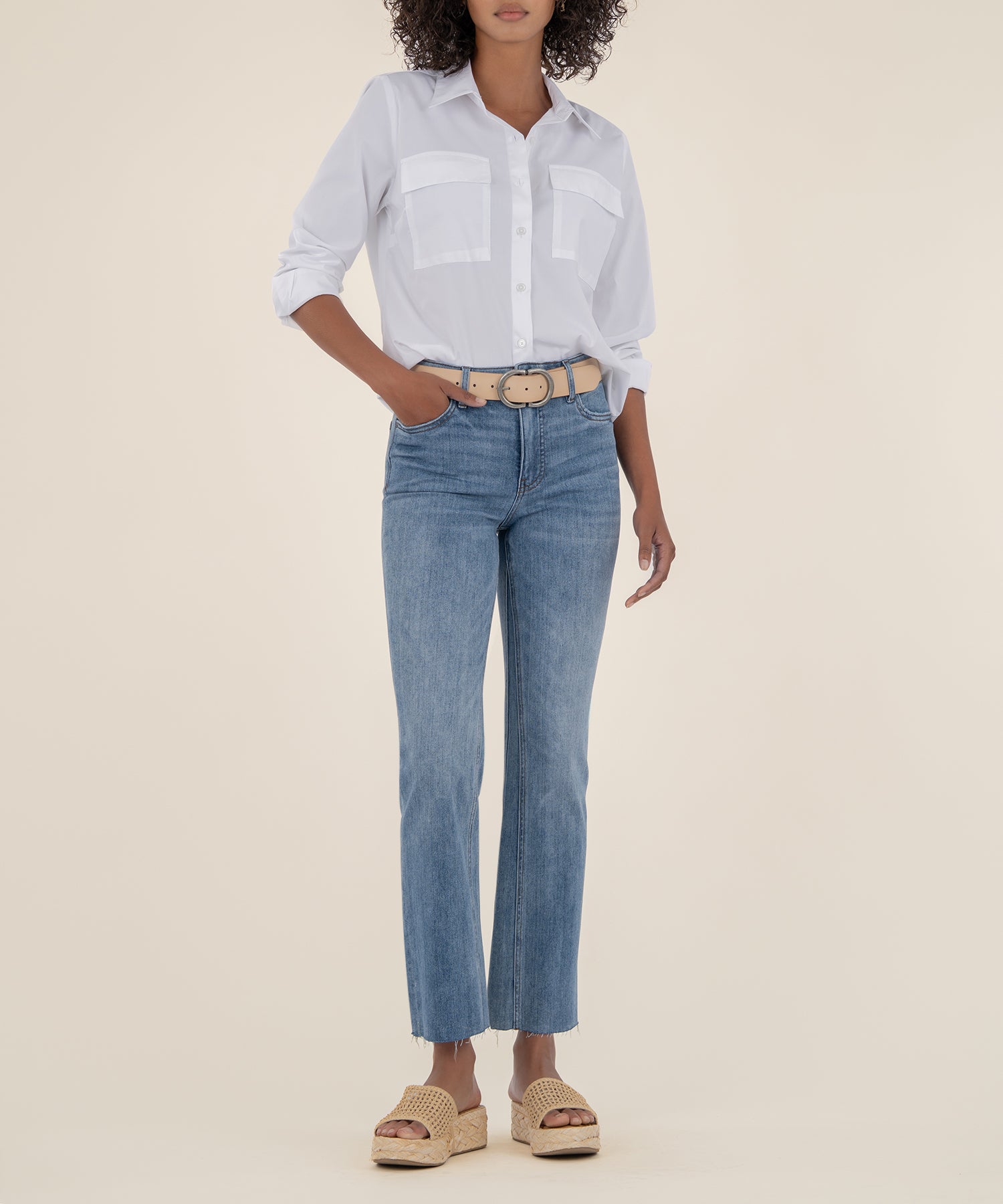 Woman wearing light blue high-rise ankle flare jeans with white button-up shirt and tan belt.