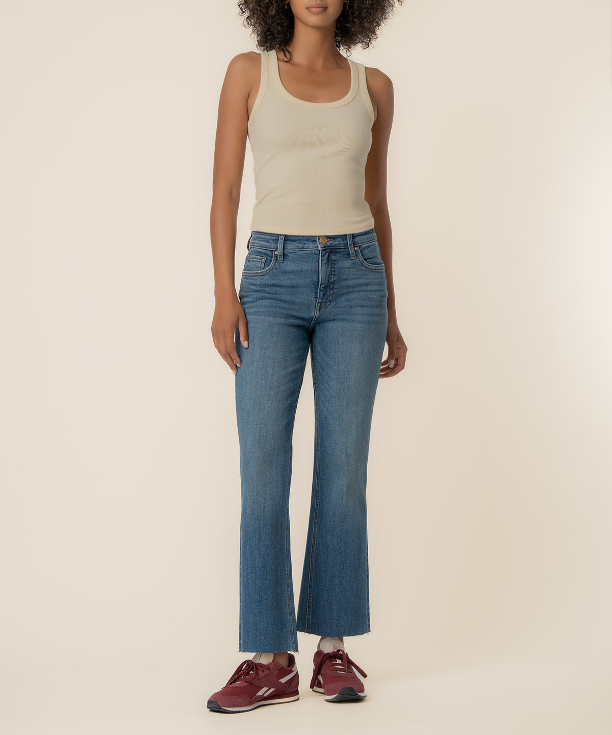 Woman wearing high-rise medium-wash ankle flare jeans with cream tank top and burgundy sneakers.