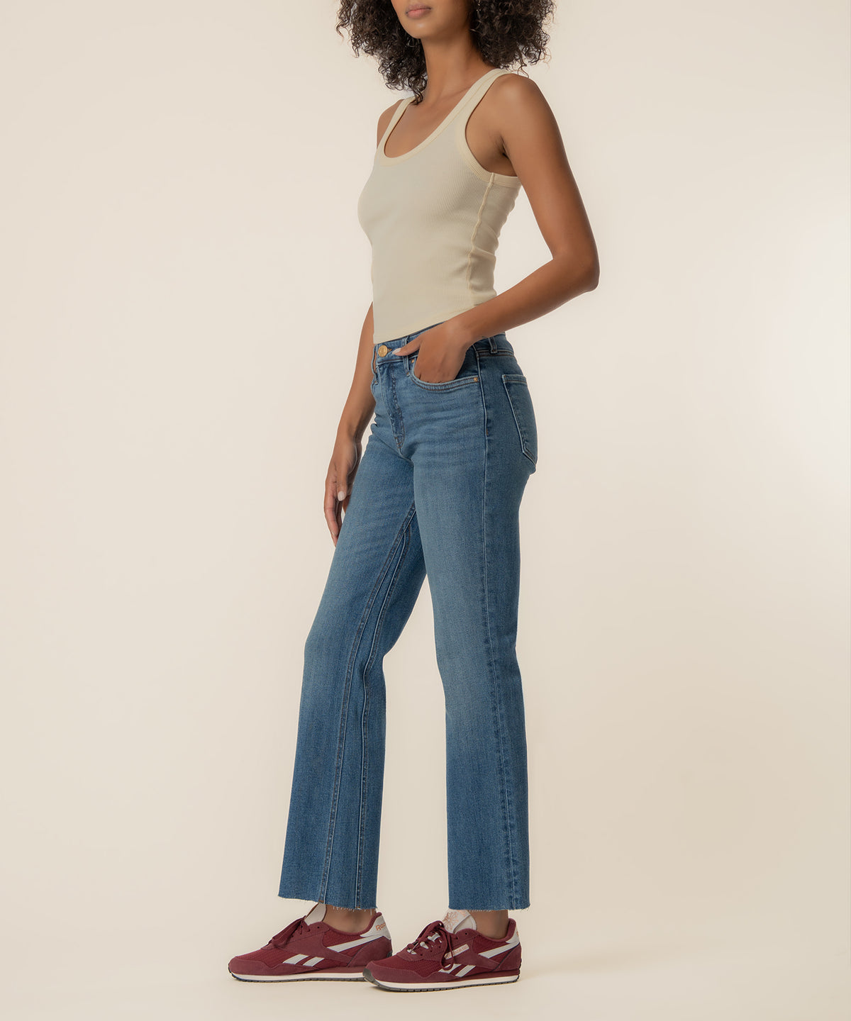 Woman wearing high-rise medium-wash ankle flare jeans with a cream sleeveless tank top and burgundy sneakers.