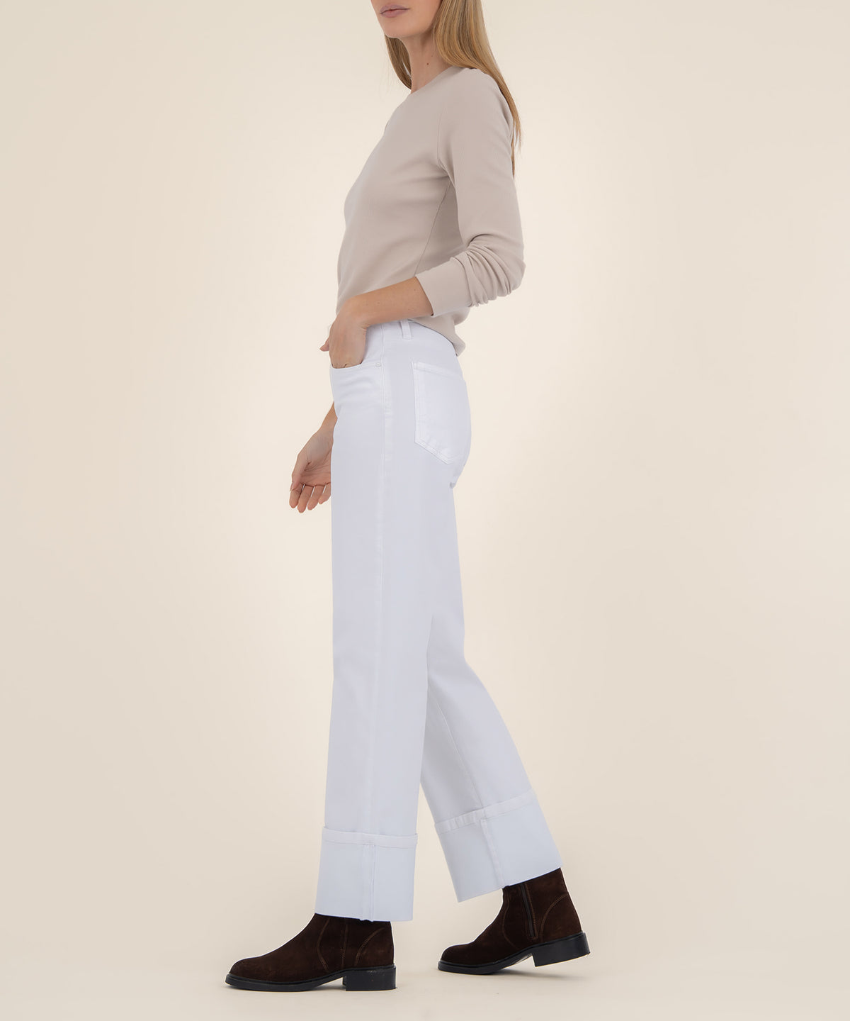 A woman in profile wears high-rise white wide-leg jeans with cuffed hems, a beige long-sleeve top, and dark brown suede boots.