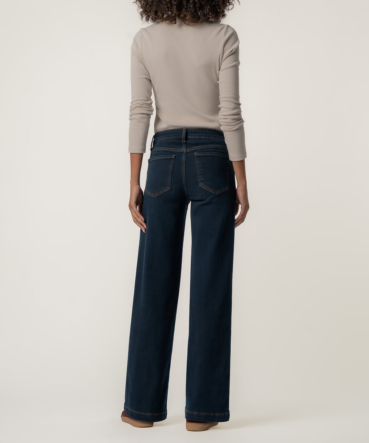 Rear view of a person wearing high-rise dark indigo wide-leg jeans with back pockets and a fitted beige long-sleeve top.