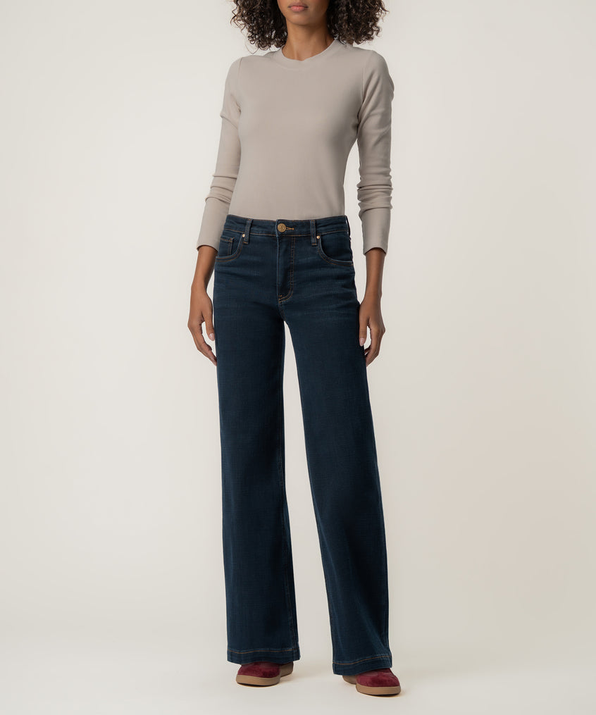 A woman stands wearing high-rise, wide straight-leg dark blue jeans with a fitted beige long-sleeve top and burgundy shoes.