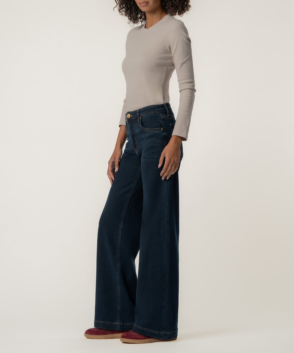 A woman stands in profile wearing dark blue high-rise wide-leg jeans, a fitted beige long-sleeve top, and burgundy shoes.
