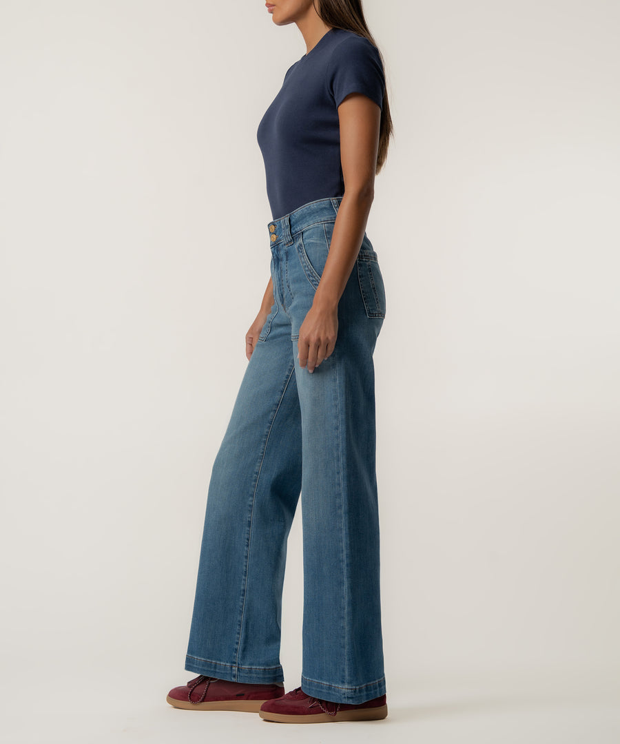 A woman stands in profile wearing high-rise, wide-leg medium-wash jeans, a navy short-sleeve top, and burgundy sneakers against a plain light background.