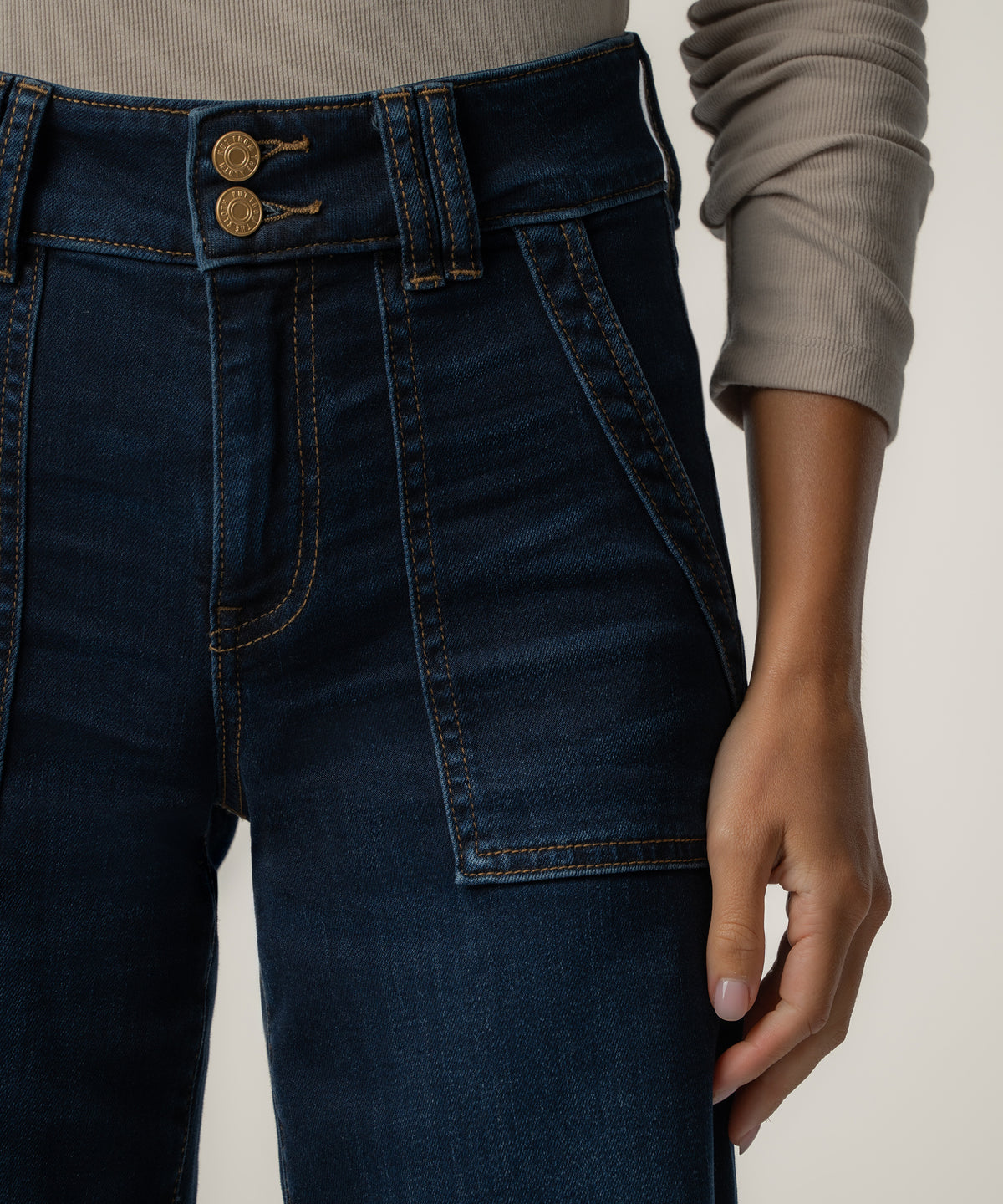Close-up of dark blue high-rise jeans with two gold buttons, orange topstitching, and a patch pocket, worn with a taupe ribbed top.