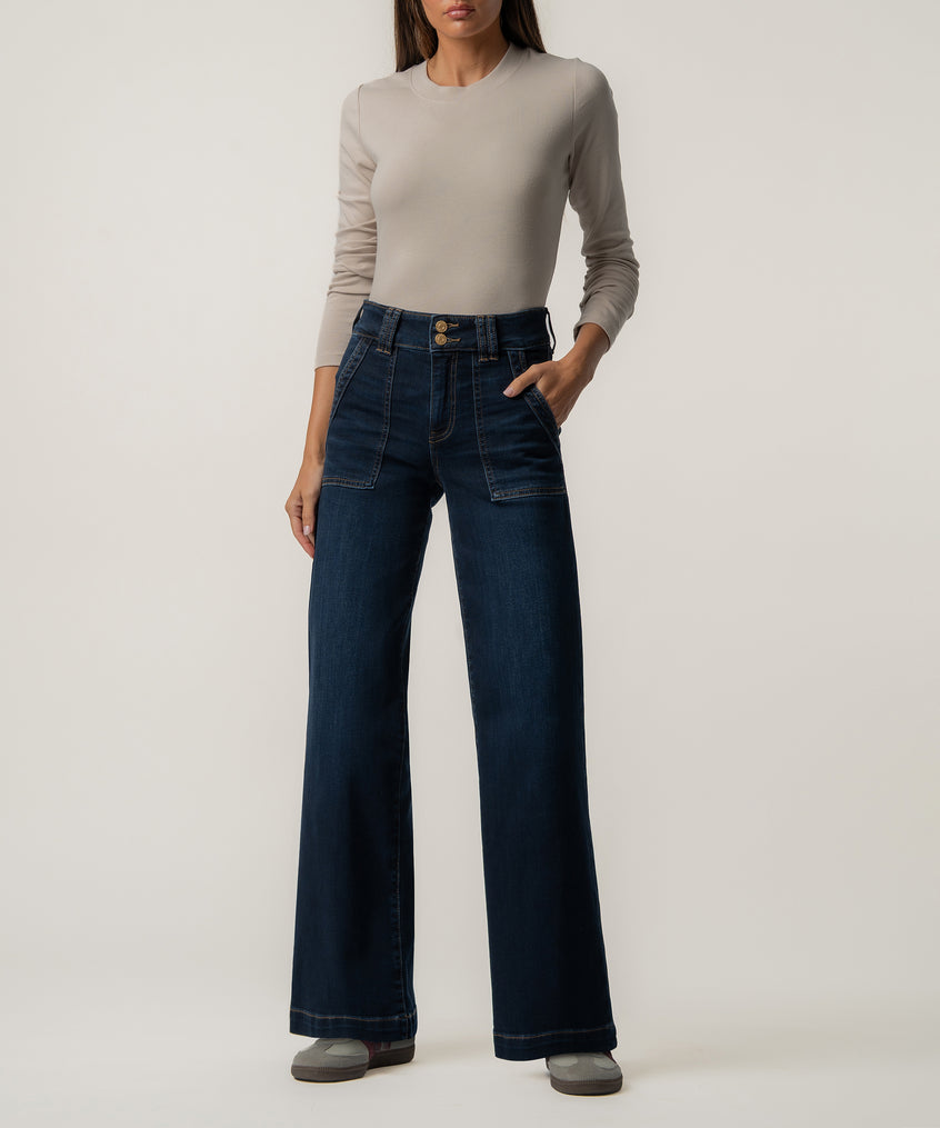 A woman wears dark blue high-rise wide-leg jeans with front patch pockets, a beige long-sleeve top, and gray sneakers.