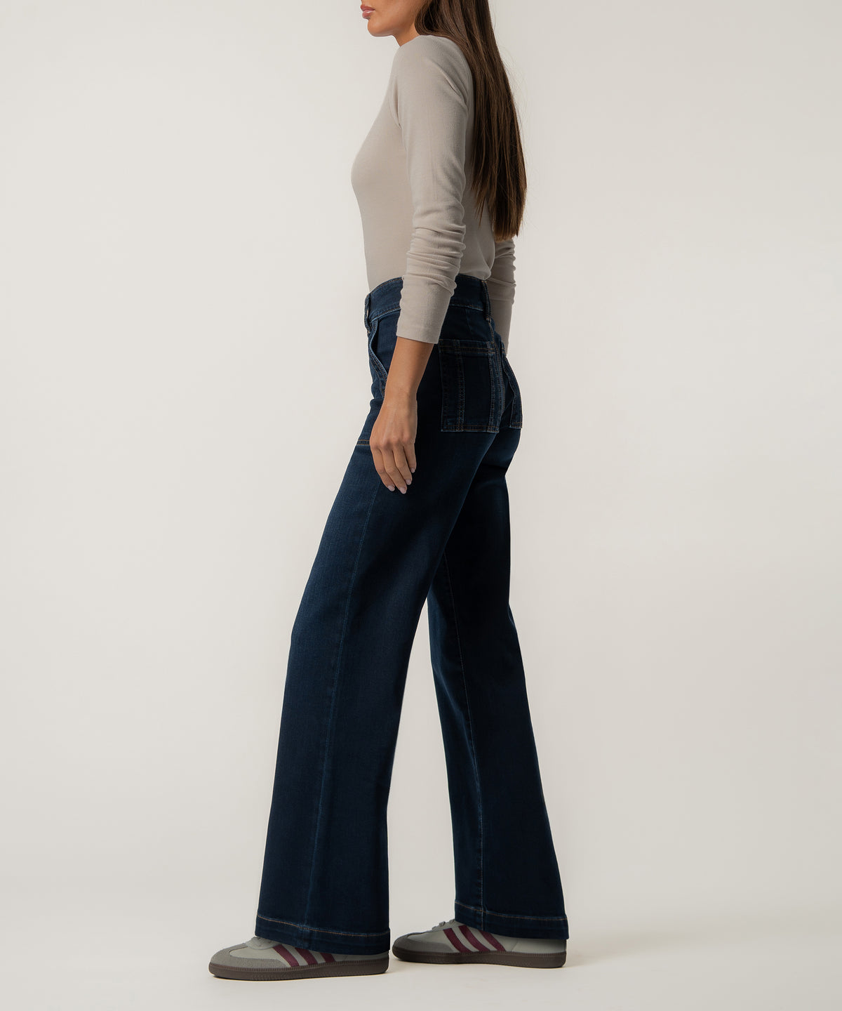 A woman stands in profile wearing dark indigo high-rise wide-leg jeans, a fitted beige long-sleeve top, and gray sneakers with maroon stripes.