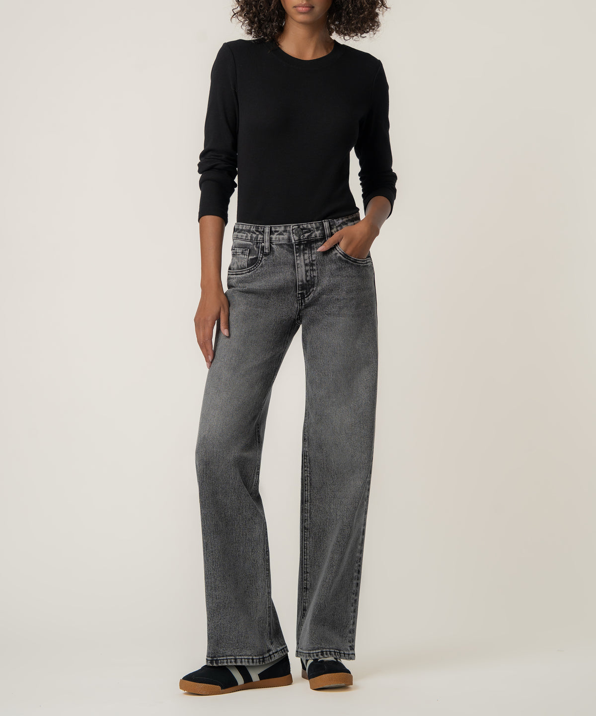 A woman stands against a neutral backdrop wearing faded gray straight-leg jeans, a black long-sleeve top, and dark sneakers with gum soles.
