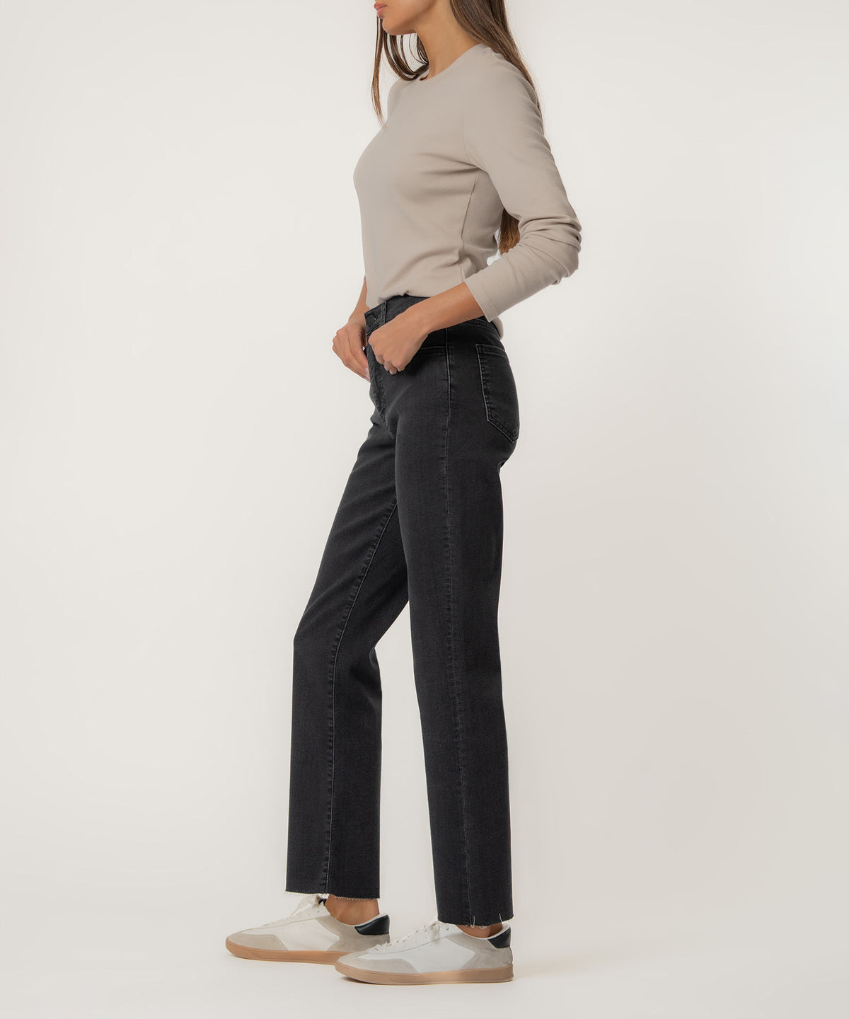 Woman wearing black high-rise straight-leg jeans with a beige long-sleeve top and white sneakers, shown from the side.