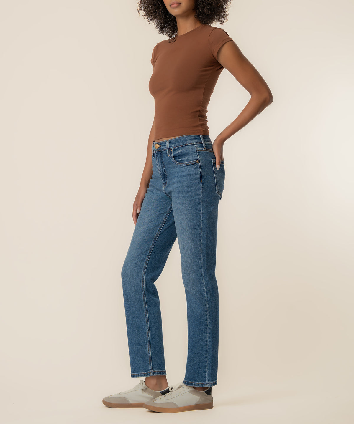 Woman wearing high-rise straight-leg blue jeans with a brown short-sleeve shirt and white sneakers.