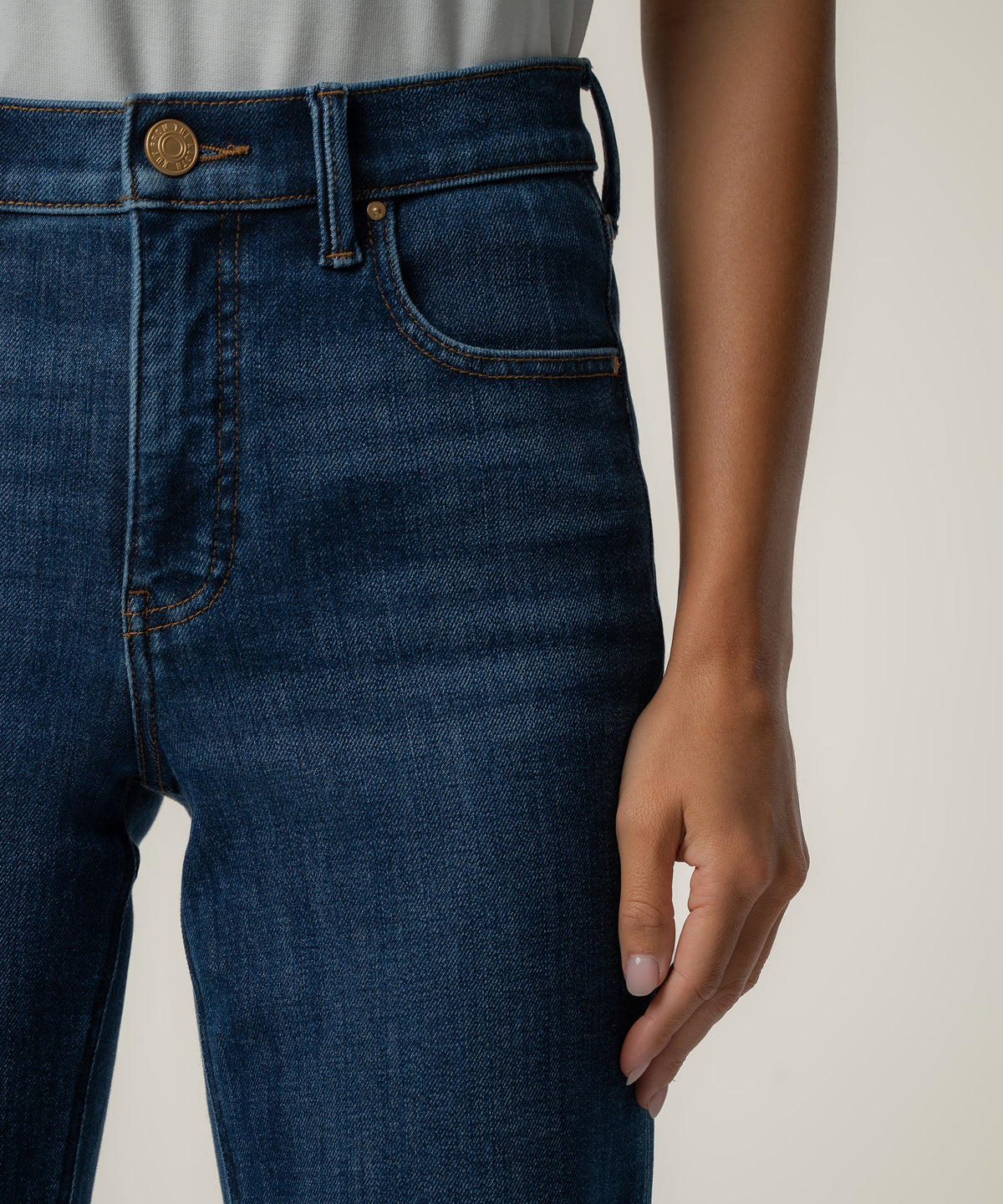 Close-up of a person wearing dark blue high-rise straight-leg jeans with a copper button and orange topstitching, paired with a light gray top.