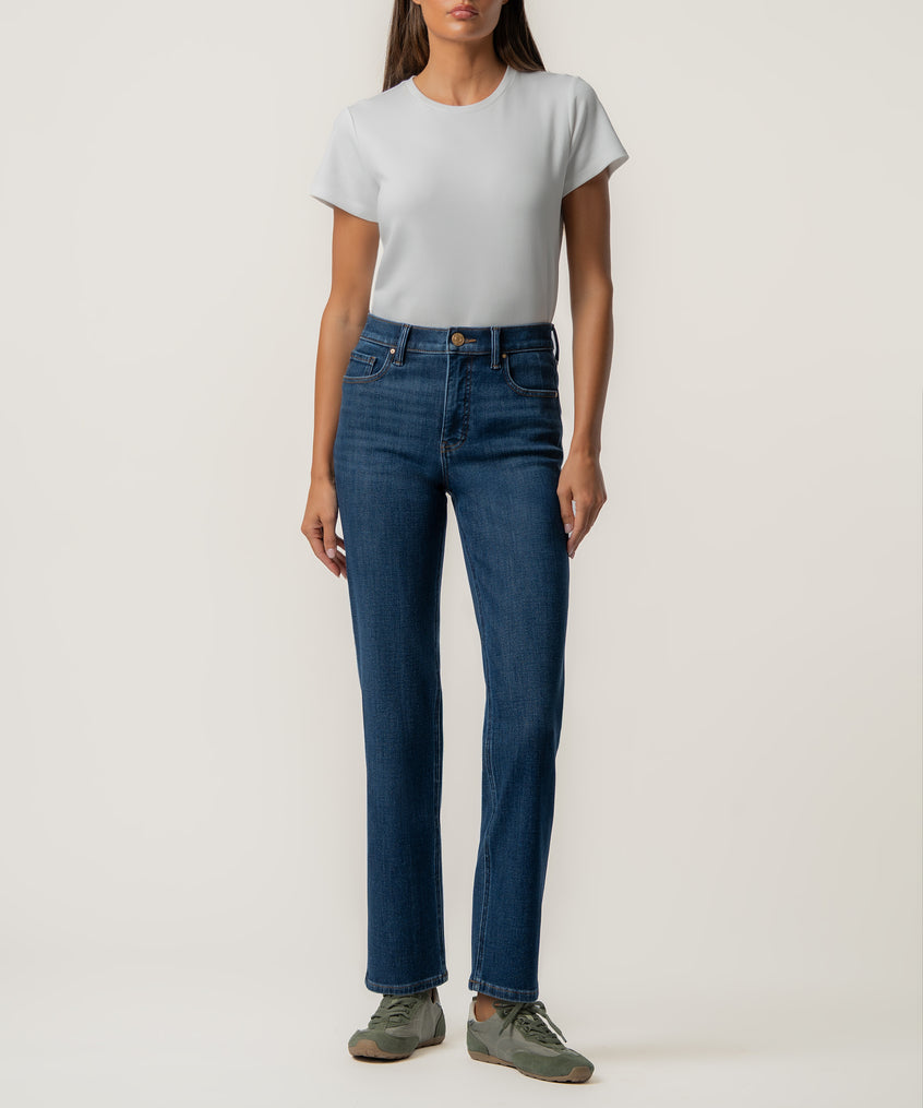 A woman stands facing forward in high-rise dark blue straight-leg jeans, a light gray short-sleeve tee, and olive green sneakers.