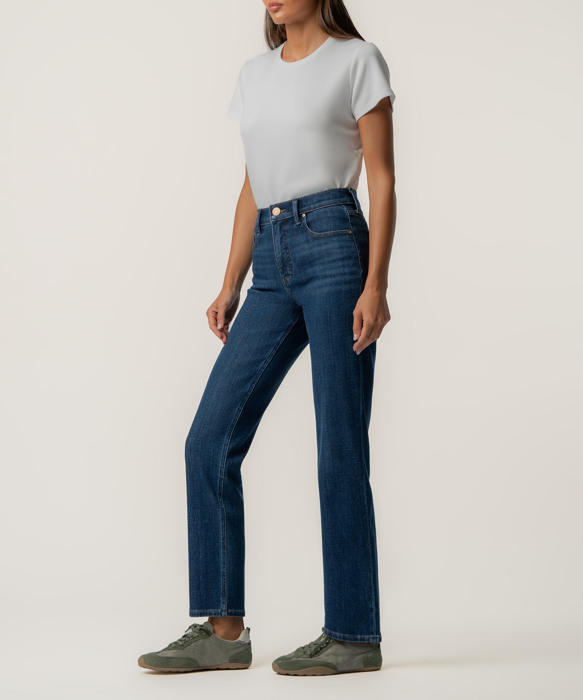A woman stands in profile wearing high-rise dark blue straight-leg jeans, a light gray short-sleeve tee, and olive green sneakers.