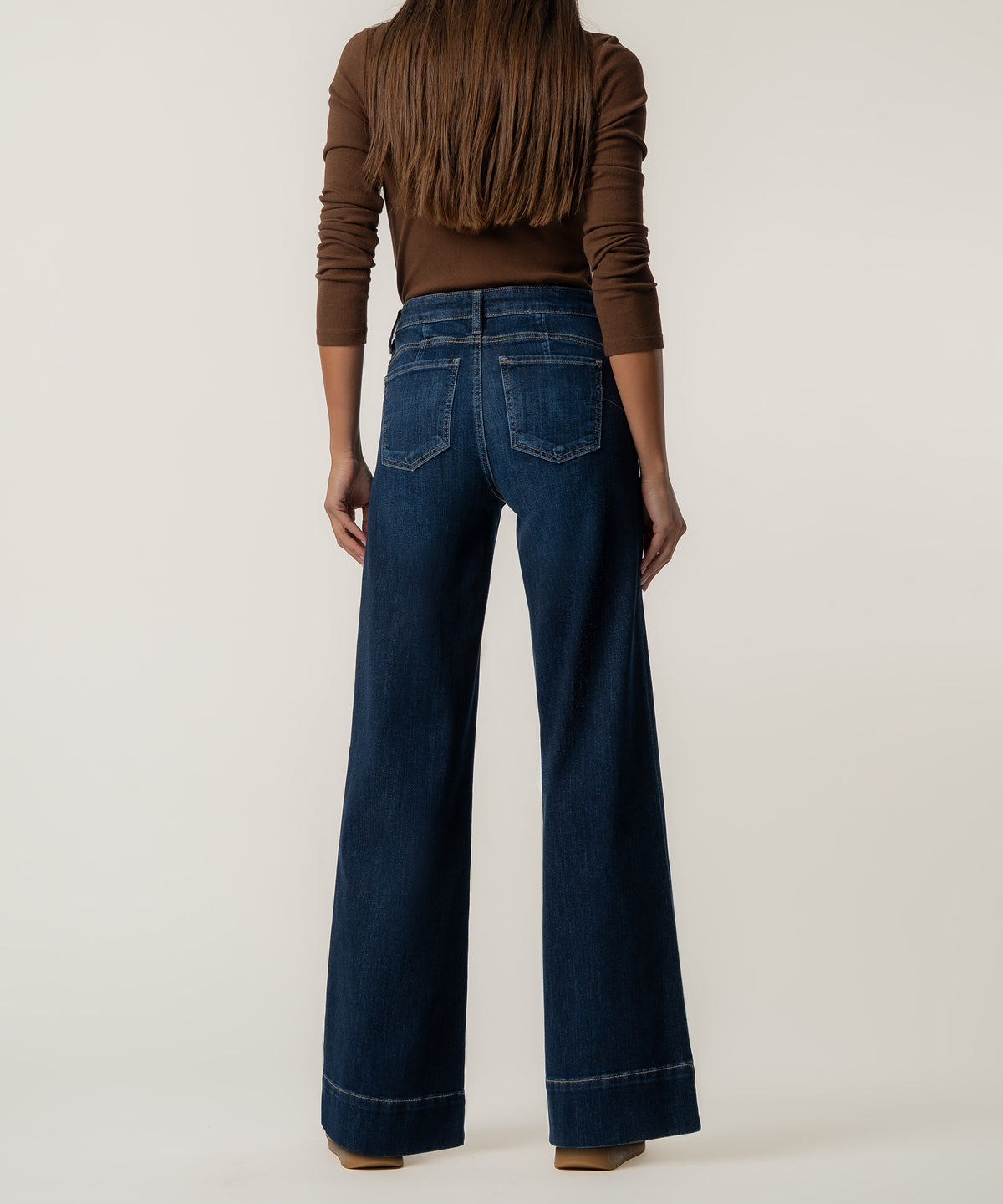 Back view of a woman wearing dark blue high-rise wide-leg jeans with back pockets, a brown long-sleeve top, and tan shoes.