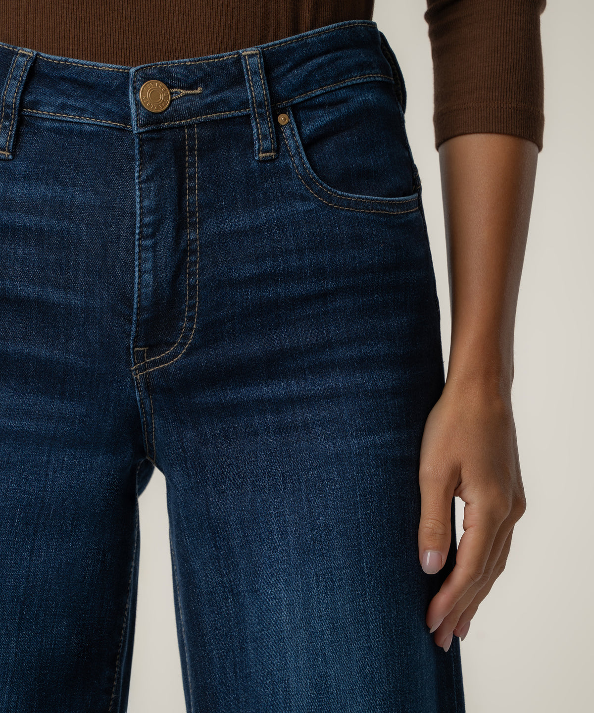 A close-up of a person wearing dark blue wide-leg jeans with a brass button and a brown long-sleeve top.
