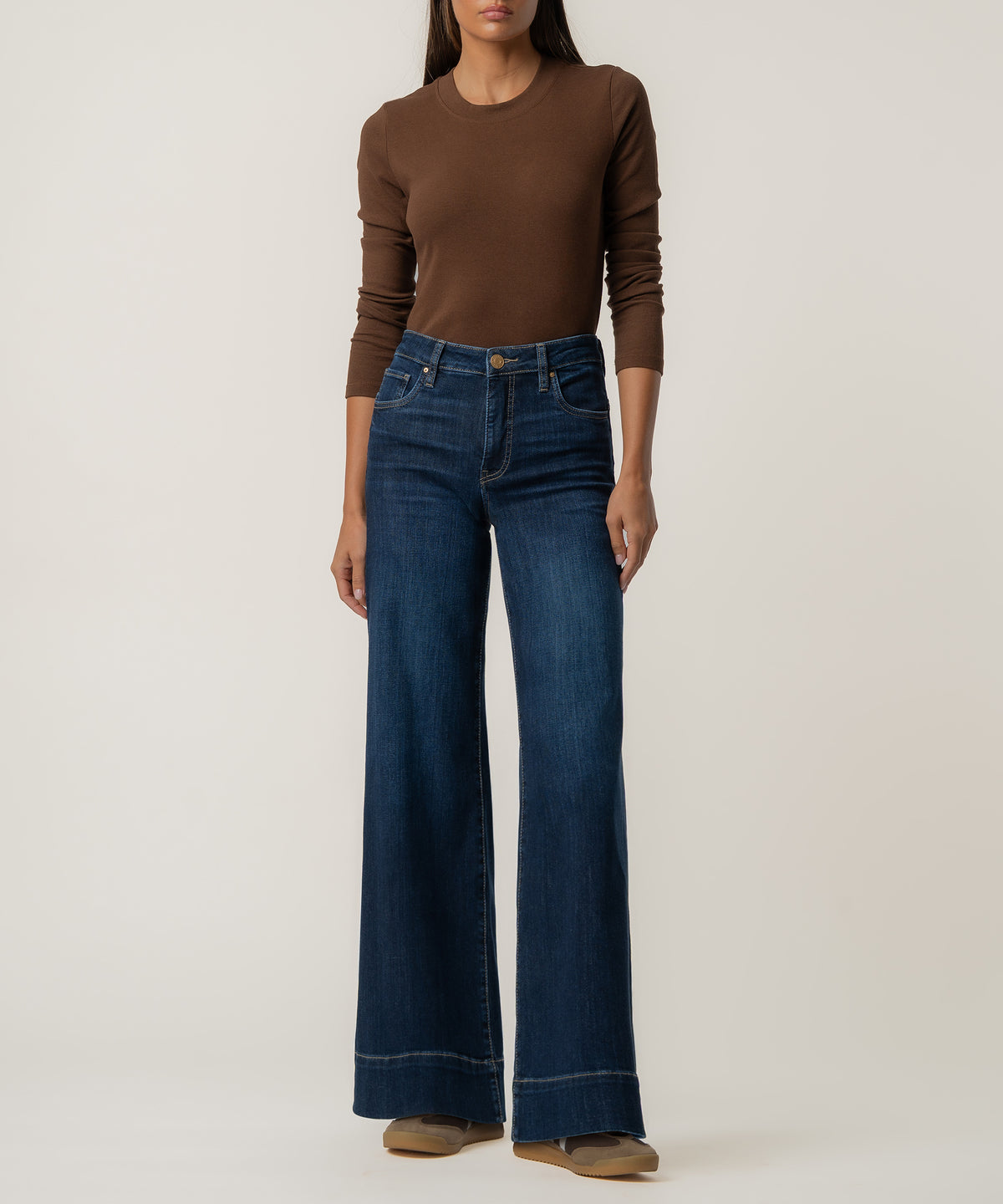 A woman stands against a plain background wearing high-rise dark blue wide-leg jeans, a fitted brown long-sleeve top, and tan shoes.