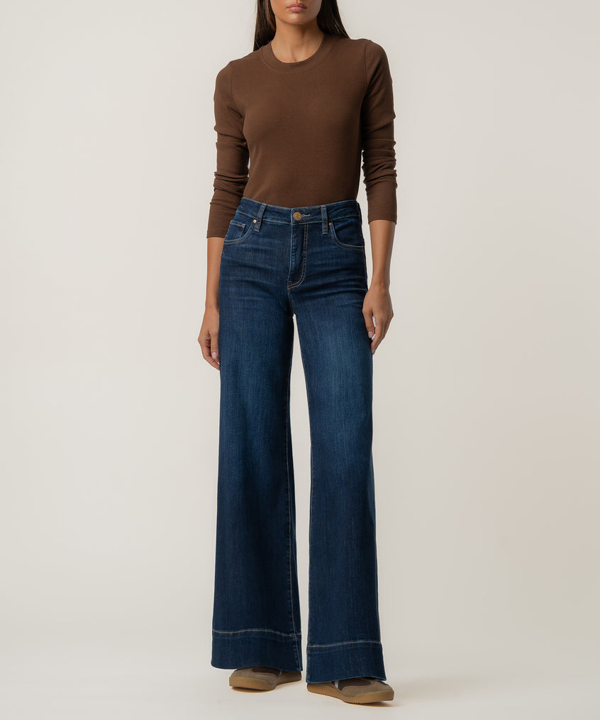 A woman stands against a plain background wearing high-rise dark blue wide-leg jeans, a fitted brown long-sleeve top, and tan shoes.